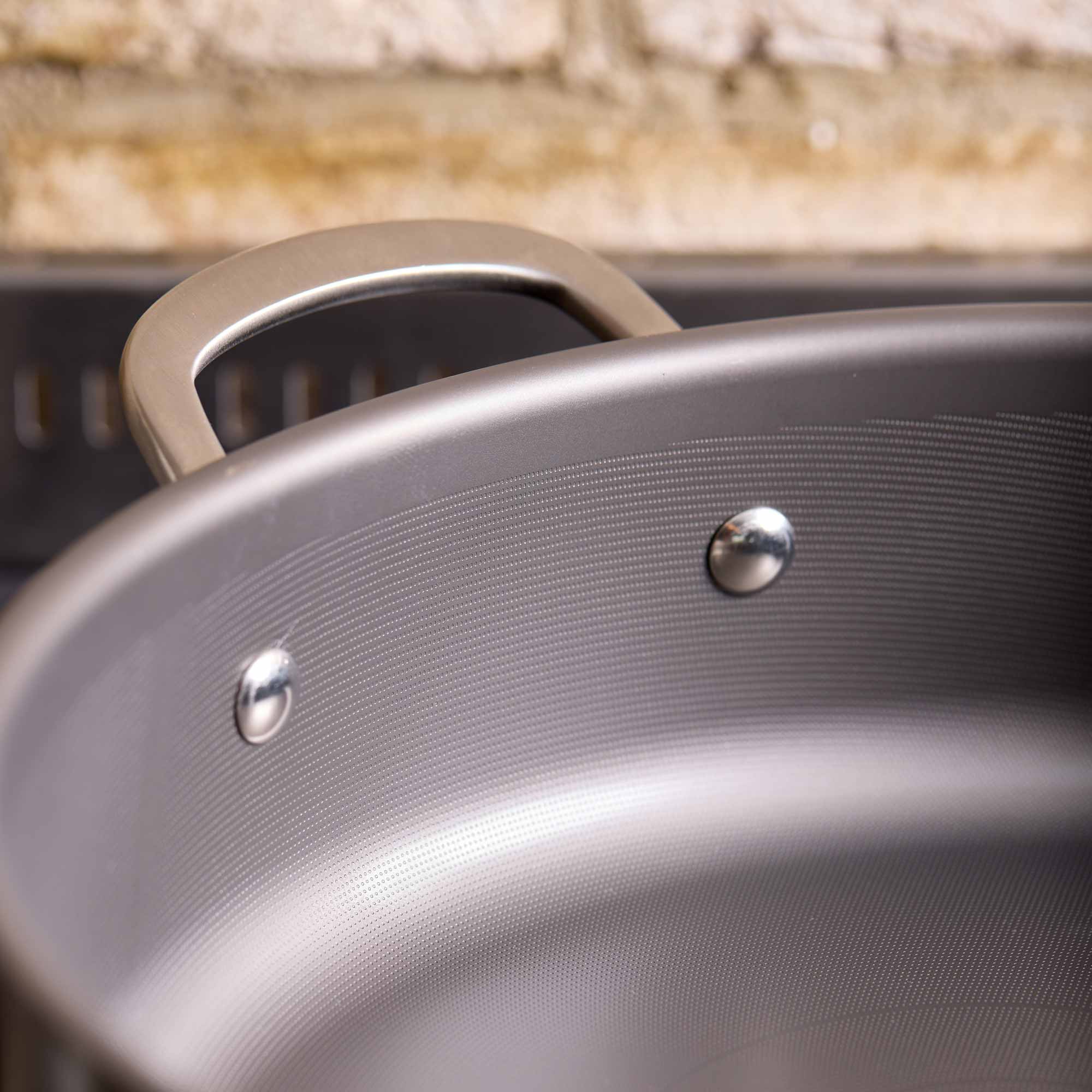 Close-up of a dark gray non-stick pan with a metallic handle and rivets, set against a blurred rustic background.