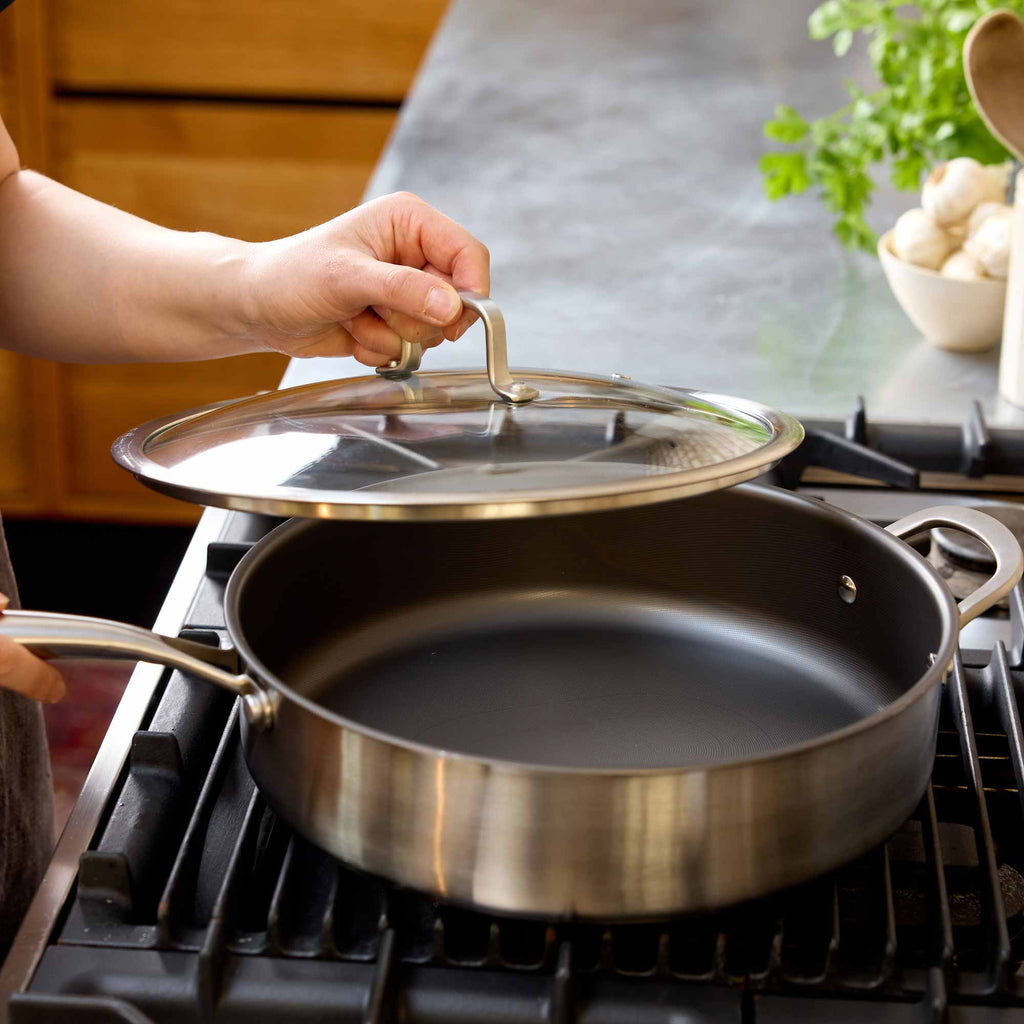 A stainless steel saute pan sits on a gas stovetop in a kitchen, with a person's hand lifting a glass lid with a metal handle.