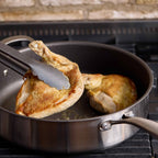A stainless steel saute pan on a stovetop with two pieces of chicken cooking in it, and metal tongs lifting one piece out of the pan.