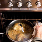 A stainless steel pot with a glass lid sits on an oven rack inside an oven, with a hand holding a beige cloth towel on the right side. The pot contains a simmering golden-brown liquid and pieces of chicken.