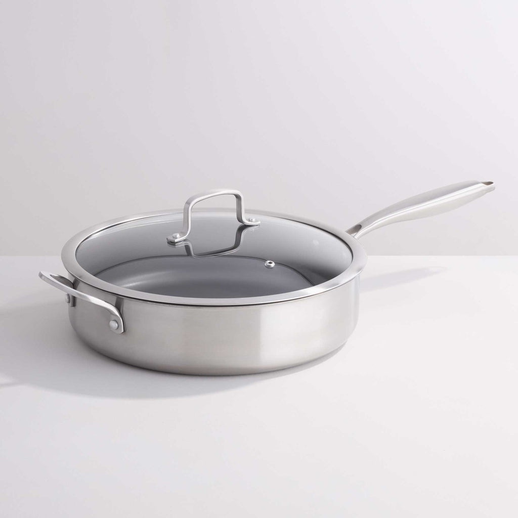 A stainless steel sauté pan with a clear glass lid and silver metal handle, photographed on a clean white studio background.