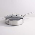A stainless steel sauté pan with a clear glass lid and silver metal handle, photographed on a clean white studio background.