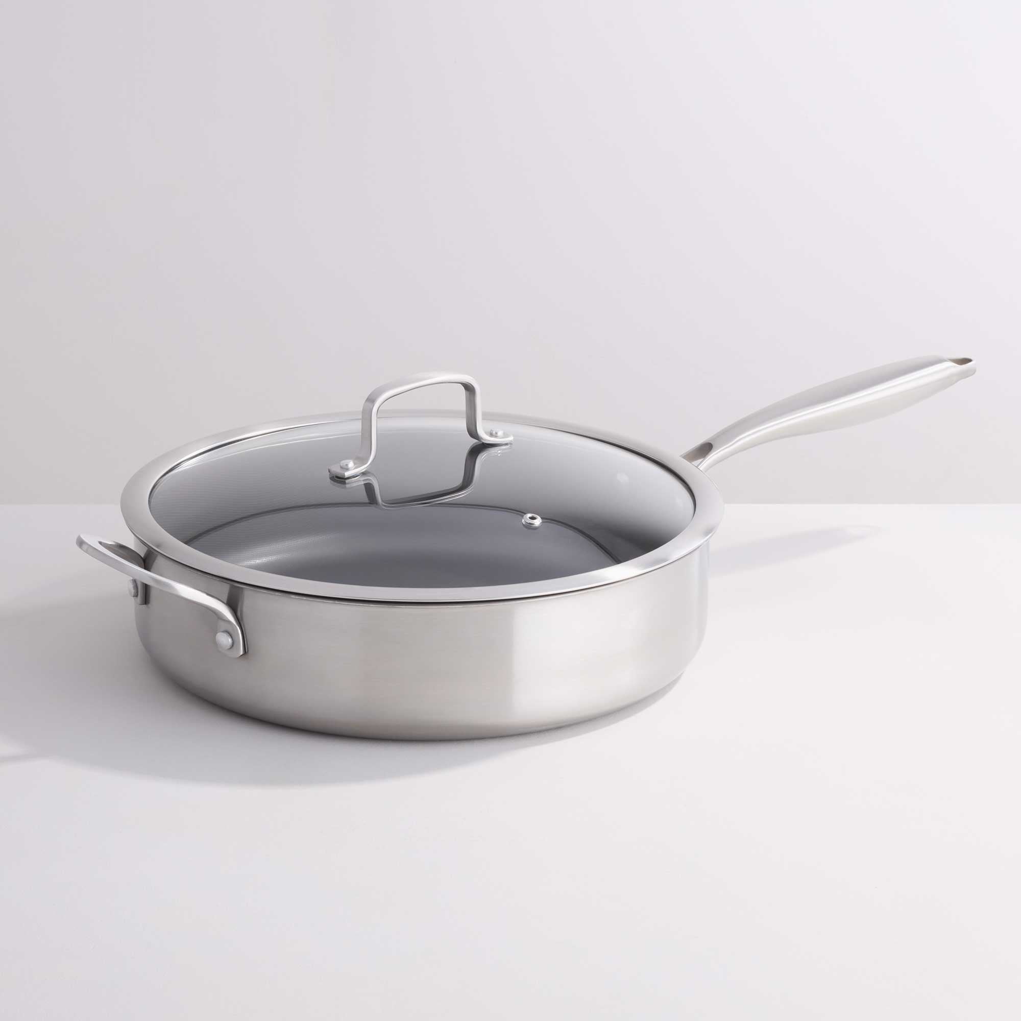 A stainless steel sauté pan with a clear glass lid and silver metal handle, photographed on a clean white studio background.