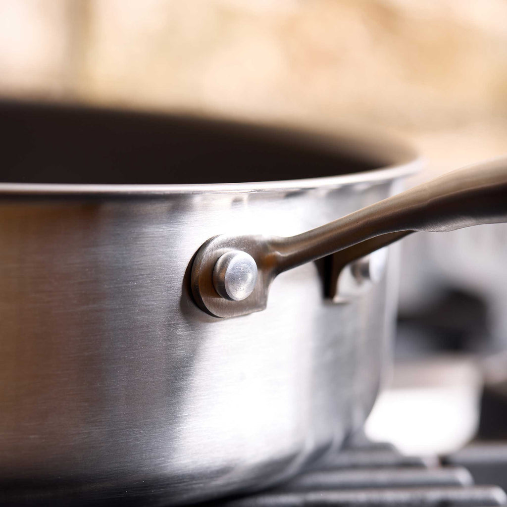 Close-up of a stainless steel saucepan with a dark metal handle and rivet, resting on a stovetop with a softly blurred kitchen background.