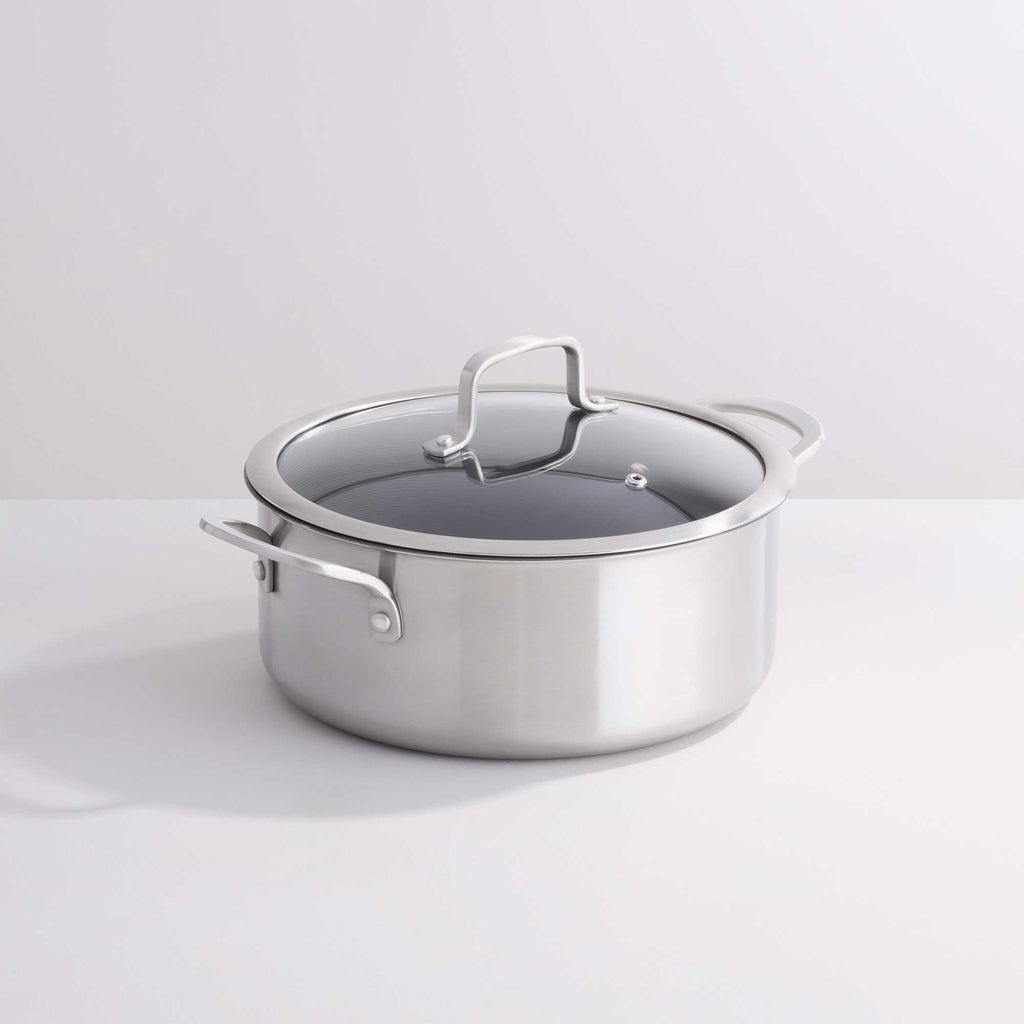 A stainless steel casserole with a glass lid and metal handles, photographed on a clean white studio background.