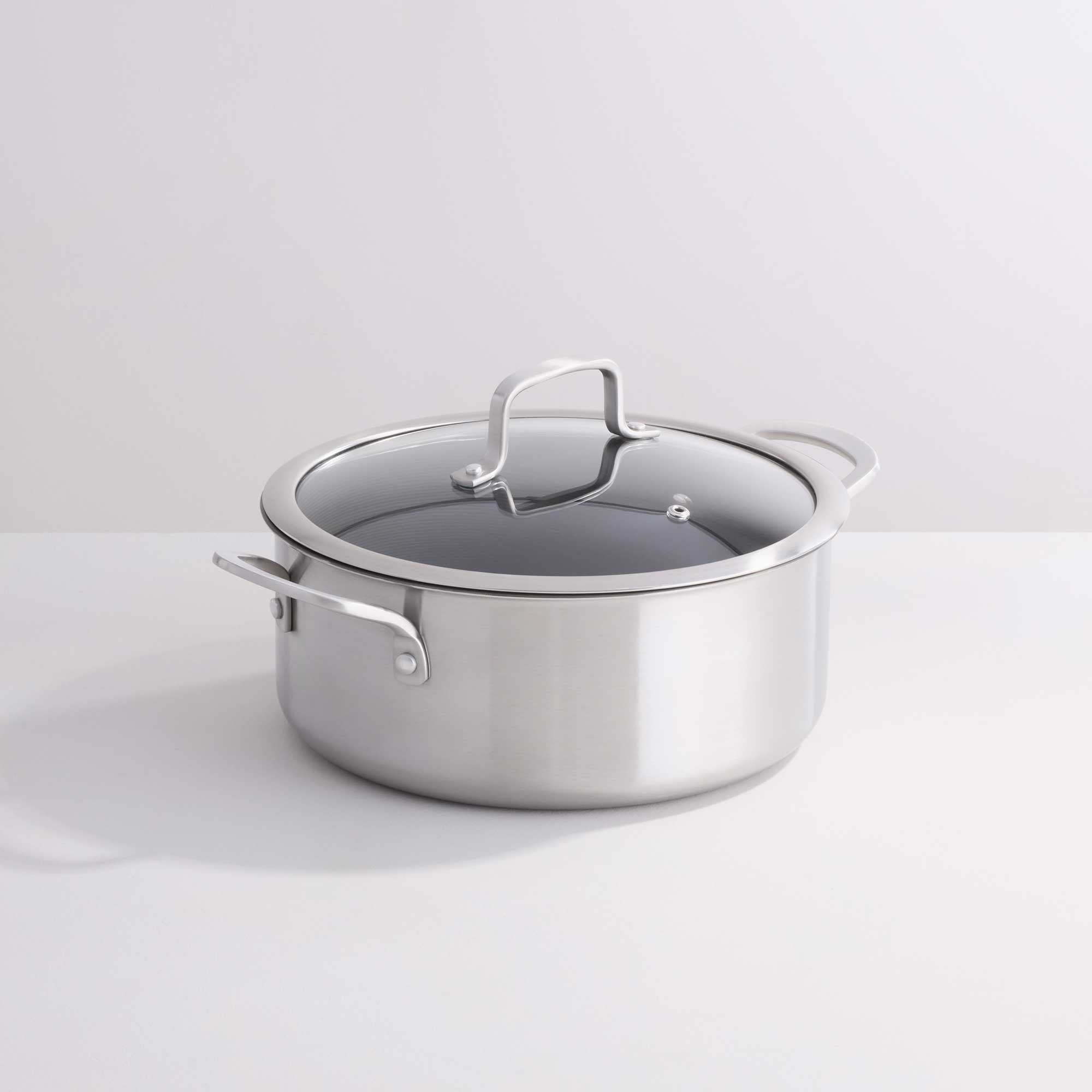 A stainless steel casserole with a glass lid and metal handles, photographed on a clean white studio background.
