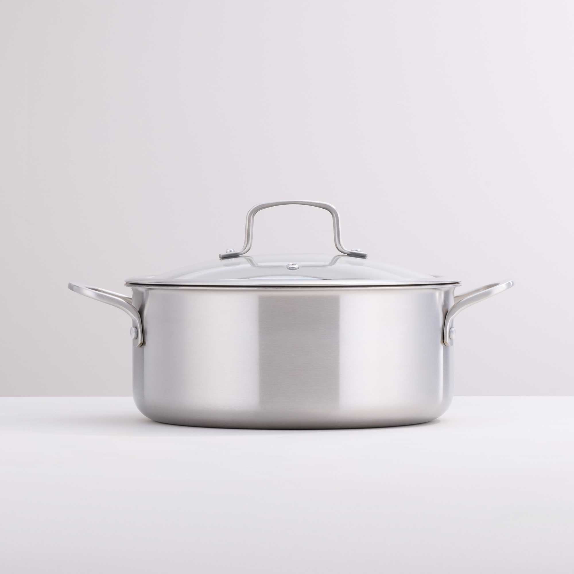 A stainless steel casserole with a glass lid and side handles, photographed on a clean white surface against a light gray background.