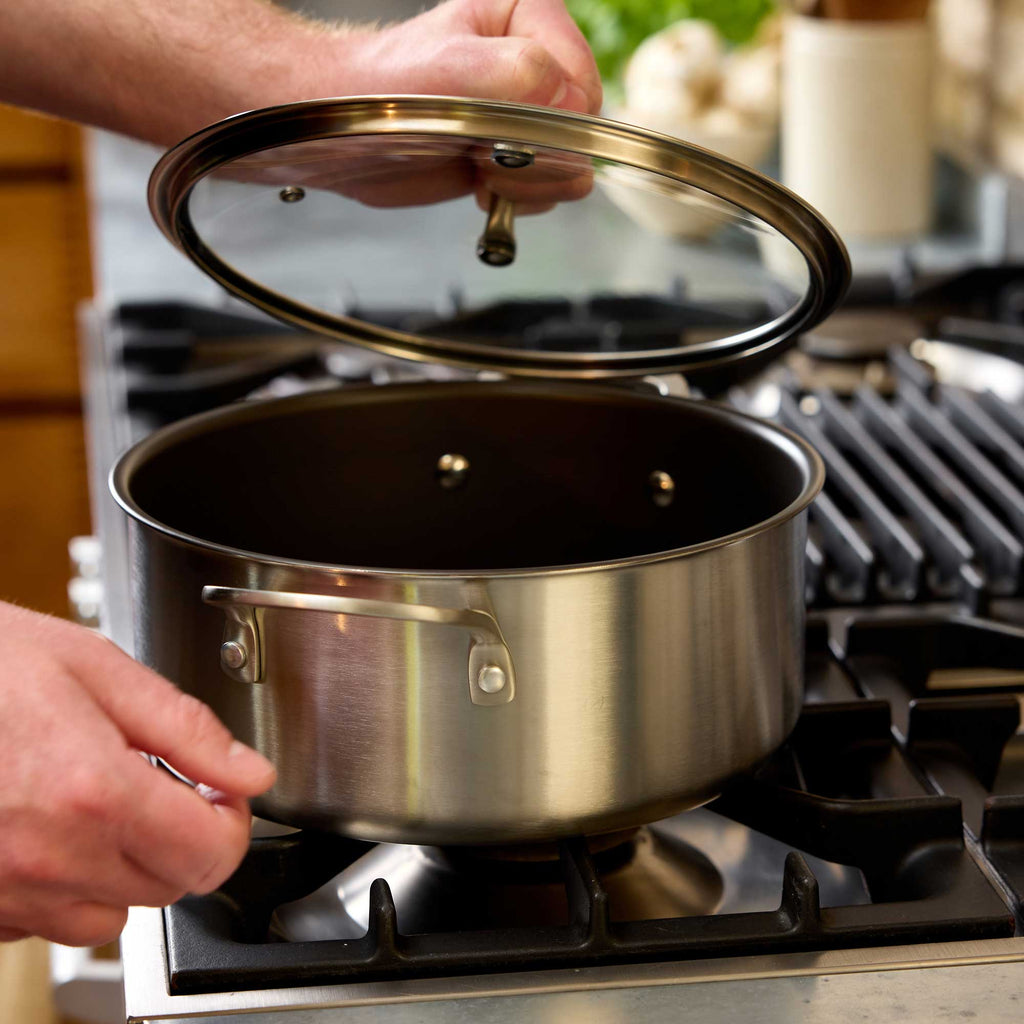 A stainless steel casserole with a glass lid being lifted by hands on a gas stovetop in a kitchen setting.
