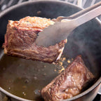 A close-up of a piece of seared beef held above a dark pot with metal tongs, with another piece cooking in the pot below.