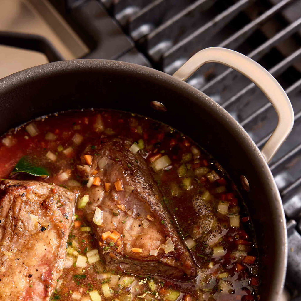 A close-up of a dark pot on a stovetop burner, containing a simmering stew with chunks of meat, onions, carrots, and herbs in a rich red-brown broth.