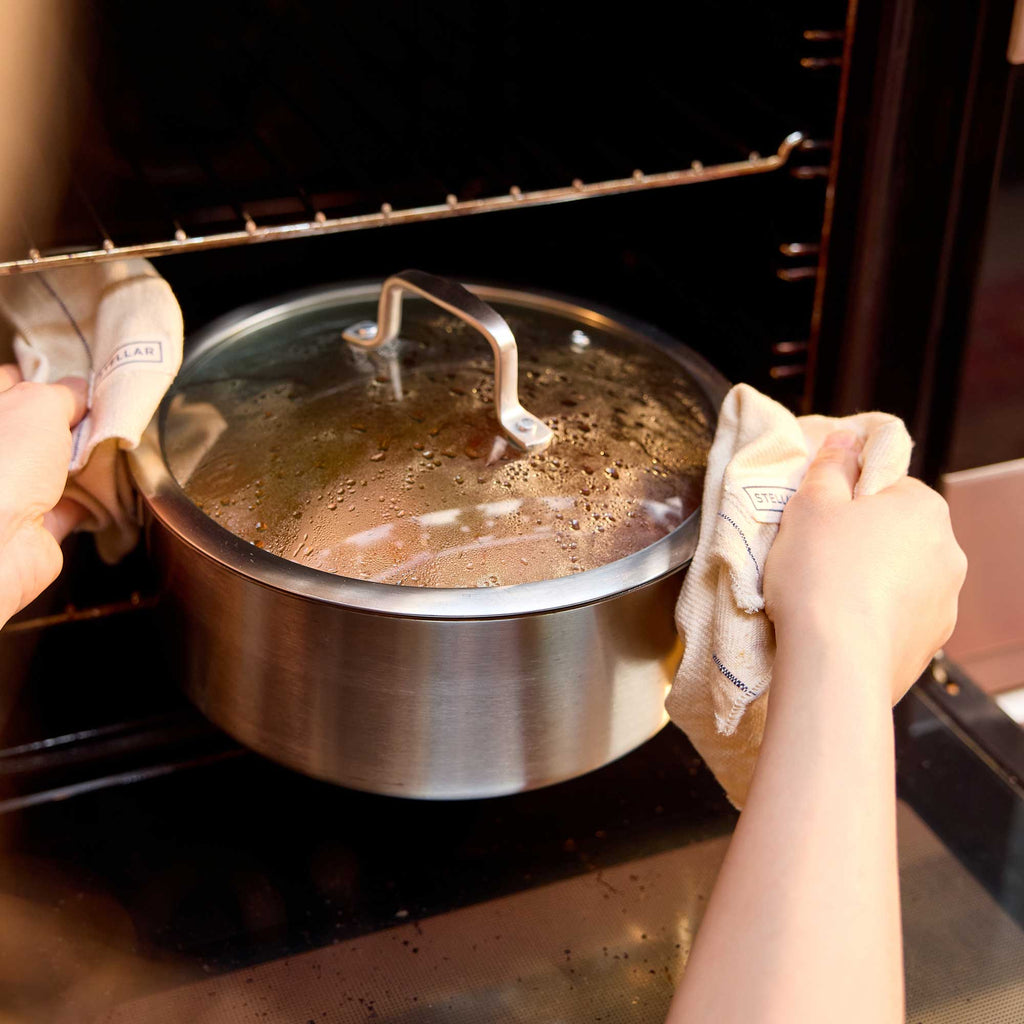 A stainless steel saucepan with a glass lid is being pulled out of an oven by two hands holding cloth towels. The pot is shown inside the oven with oven racks visible in the background.
