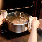 A stainless steel saucepan with a glass lid is being pulled out of an oven by two hands holding cloth towels. The pot is shown inside the oven with oven racks visible in the background.