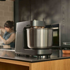 A Ooni Halo Pro Spiral Mixer in Charcoal Grey on a kitchen counter with someone baking in the background.