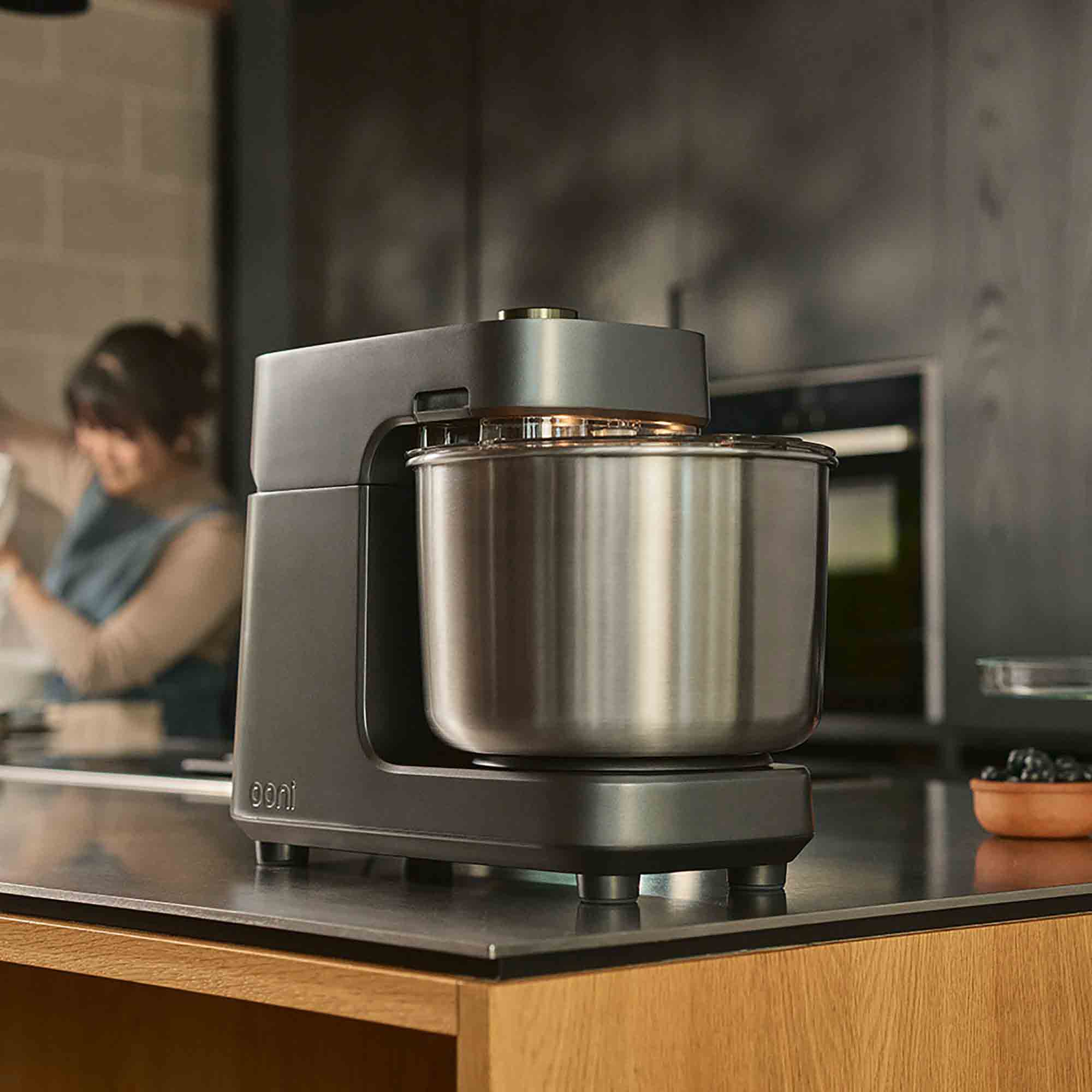 A Ooni Halo Pro Spiral Mixer in Charcoal Grey on a kitchen counter with someone baking in the background.