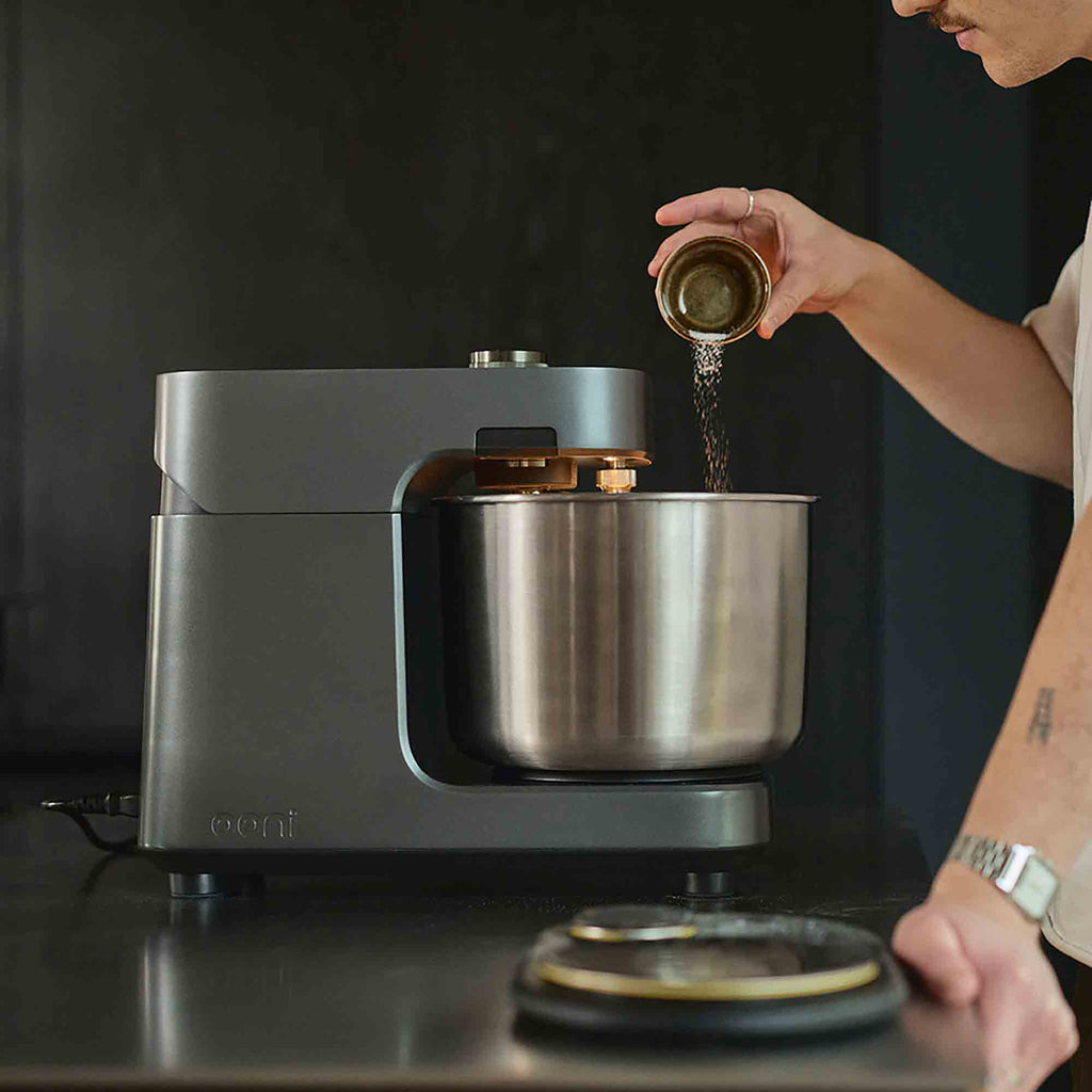 Someone adding ingredients to the Ooni Spiral Mixer on a kitchen counter.