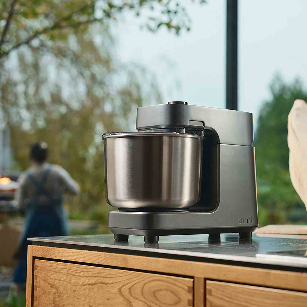 A Ooni Spiral Mixer in Charcoal Grey on a kitchen island counter with someone baking in the background.