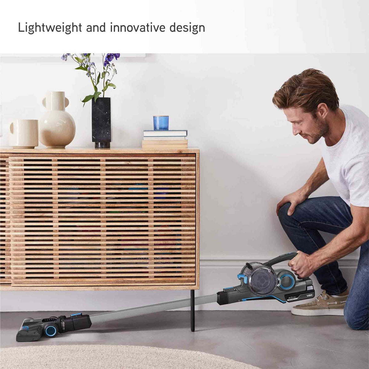 A man kneels on the floor using a cordless stick vacuum cleaner in a modern living room setting, with a wooden slat cabinet and decorative items in the background. Text at the top reads 'Lightweight and innovative design'.